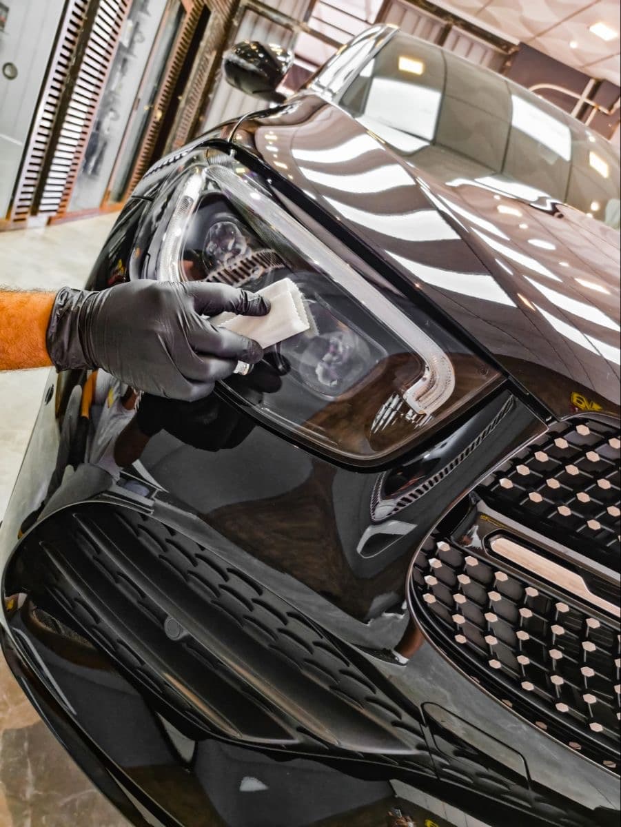 Gloved hand applying a protective coating to the headlight of a sleek black car.
